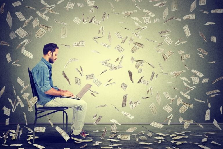 Young man using a laptop building online business making dollar bills cash falling down. Beginner IT entrepreneur under money rain